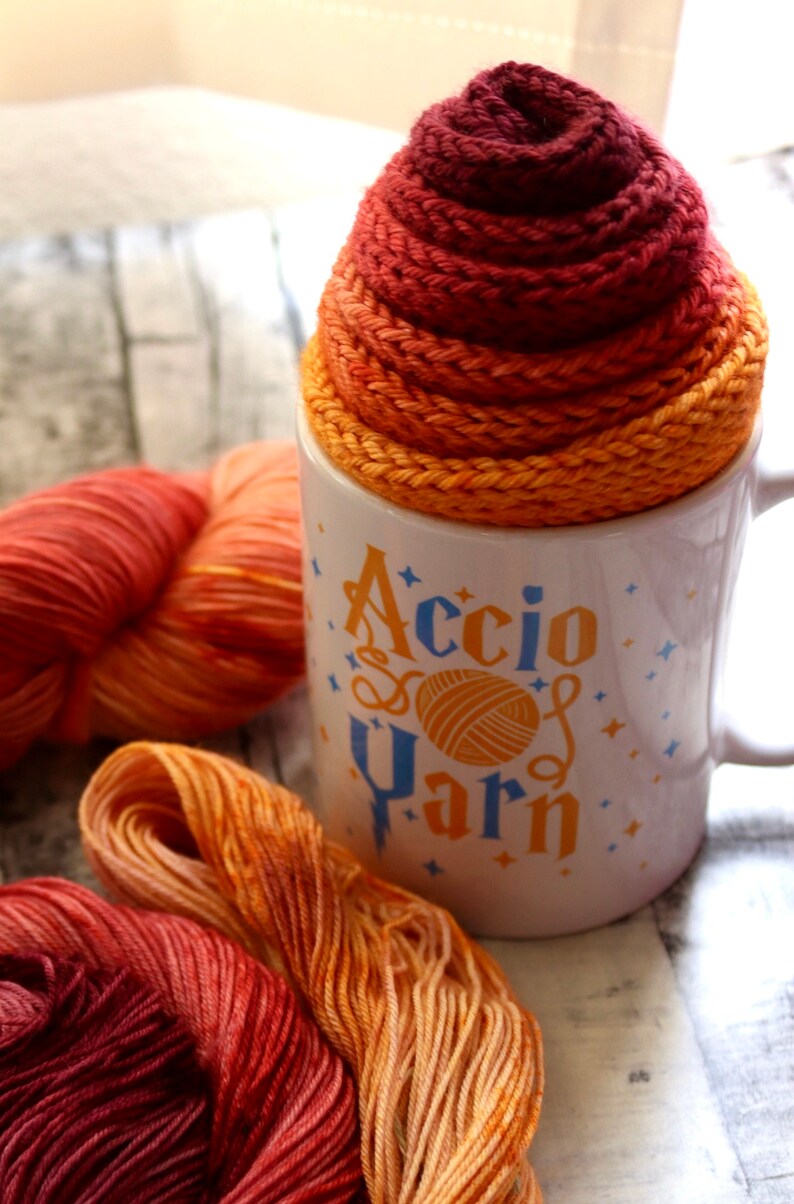 Peut inclure: Une tasse en c&eacute;ramique blanche avec le texte "Accio Yarn" en lettres bleues et jaunes. La tasse est remplie d'une pelote de laine dans des tons d'orange, de jaune et de rouge.