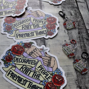 May include: A collection of stickers and keychains with a knitting theme. The stickers and keychains feature a banner with the text "Decorated People Knit the Prettiest Things" and illustrations of hands knitting with red roses.