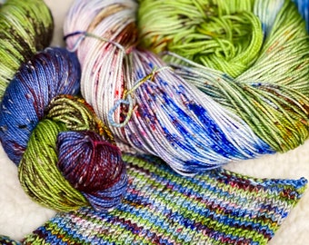 May include: Three skeins of yarn in shades of green, blue, and purple. The yarn is hand-dyed and has a speckled appearance. A knitted swatch of the yarn is also shown.