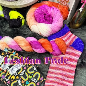 Op de afbeelding: Een set kleurrijke garenklossen in tinten roze, oranje en wit, met een regenboog gestreepte sok en een stoffen tas met een regenboog en hartpatroon. De stoffen tas heeft de tekst "Lesbian Pride" erop.