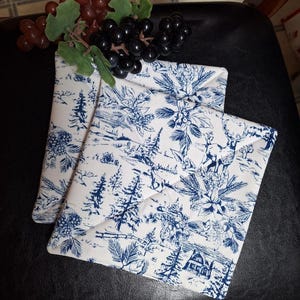 May include: Two square fabric pot holders featuring a white background with a blue toile-style print depicting winter scenes, including pine trees, a cabin, a deer, and holly berries, topped with a cluster of artificial red and black grapes.