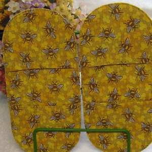 May include: A pair of yellow oven mitts with a bee and honeycomb pattern. The mitts are made of fabric and have a quilted design.