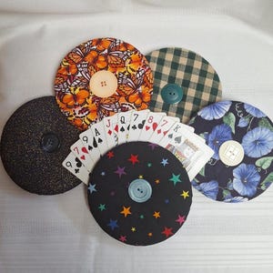 May include: Six fabric coasters with diverse patterns, such as butterflies, gingham, floral, and stars. Each coaster features a central button. A deck of playing cards is fanned out in the middle of the coasters.