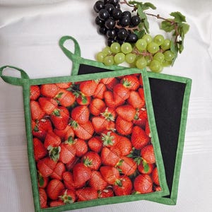 May include: Two square pot holders. One has a strawberry print with a green border and loop. The other is solid black with a green border. Grapes are in the background.
