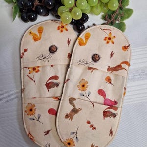 Peut inclure: Une paire de maniques beiges avec un motif de fleurs et d'écureuils. Les maniques ont une poche et sont décorées d'accents orange, marron et rouge. Le fond est beige clair.