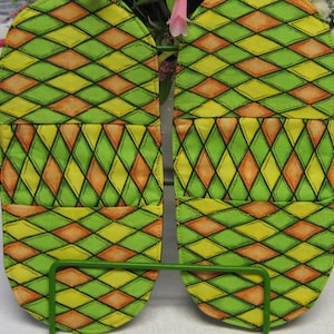 May include: A pair of green, yellow and orange fabric pot holders with a diamond pattern. The pot holders are hanging on a metal rack.