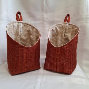 May include: Two hanging storage baskets made of a woven fabric in a rust orange colour. The baskets have a cream coloured fabric lining with a subtle pattern. The baskets have a loop at the top for hanging.