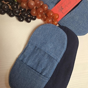 Peut inclure: Deux maniques ovales en tissu denim, une bleu clair et une bleu marine foncé, dotées d'une poche horizontale au centre. Une bande de papier rouge avec le texte manuscrit Kady est enroulée autour de la manique bleu clair.
