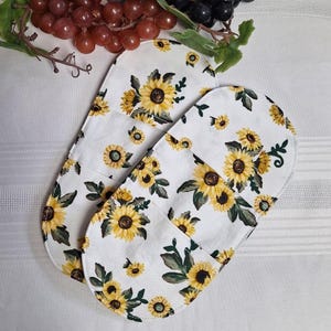 Peut inclure: Deux maniques blanches avec un motif de tournesols. Les maniques présentent un motif répété de tournesols jaunes avec des feuilles vertes. Le fond est blanc. Les maniques sont de forme ovale et semblent être en tissu.