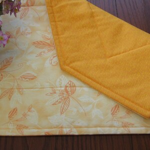 May include: A yellow table runner with a white floral pattern. The runner is folded in half and has a quilted texture.