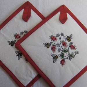Peut inclure: Deux maniques en coton blanc avec une bordure rouge et des fraises et des abeilles brodées.