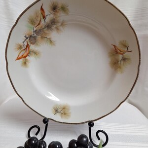 Peut inclure: Assiette en céramique blanche avec un bord festonné et un bord marron. L'assiette présente un motif peint d'aiguilles de pin, de pommes de pin et d'un petit oiseau dans des tons orange, vert et marron.