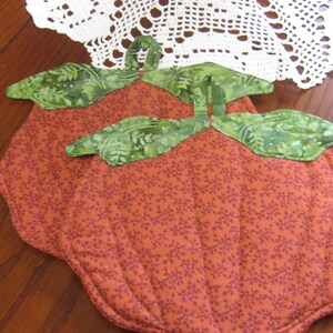 Peut inclure: Deux sets de table matelassés en forme de citrouille. Les sets de table sont orange avec un petit motif floral et des accents de feuilles vertes. Les sets de table sont sur une table en bois avec une nappe en dentelle blanche en arrière-plan.