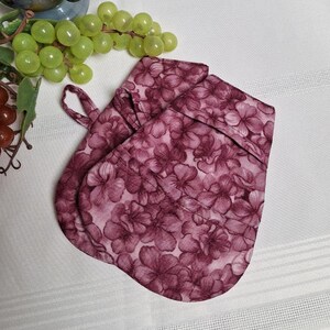 Peut inclure: Une paire de maniques de cuisine en tissu matelassé rose à fleurs avec une poignée en boucle. Les maniques sont imprimées de fleurs roses et blanches.
