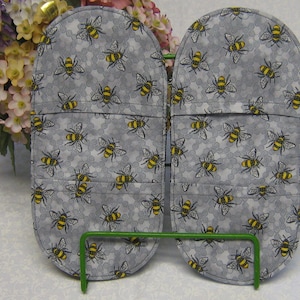 May include: Two grey oven mitts with a bee pattern. The mitts are on a green metal stand.