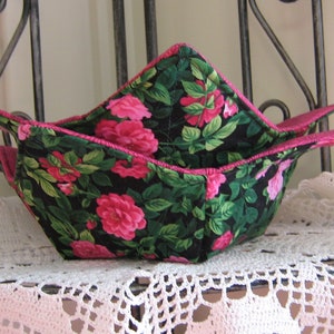 May include: Two fabric bowl cozies with a floral pattern of pink roses and green leaves on a black background. The edges are trimmed with pink fabric. The cozies are sitting on a white lace doily.