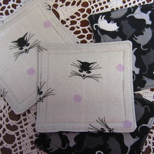 May include: Four square fabric coasters featuring a cat design. The coasters are white with black cat silhouettes and pink polka dots. One coaster has a black and white cat design with multiple cats.