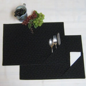 May include: Two black quilted placemats with a folded pocket to hold cutlery. The placemats are on a white surface with a small jug and grapes.