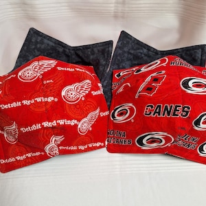 May include: Two bowl cozies. One is red with white Detroit Red Wings logos and text. The other is red with black Carolina Hurricanes logos and text. Both have a dark grey backing.