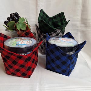 Peut inclure: Quatre manchons de bol en tissu à carreaux rouges, bleus, verts et gris, chacun contenant un pot de crème glacée. Les manchons ont une base carrée et des côtés pliés. Un petit vase décoratif de raisins est en arrière-plan.