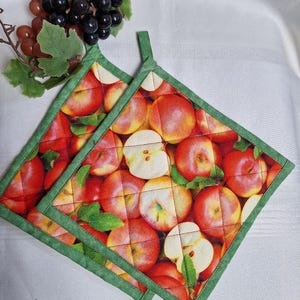 May include: Two square quilted pot holders featuring a vibrant print of whole and sliced red apples with green leaves. Each pot holder is finished with a solid green fabric border and includes a small fabric loop at the top corner for hanging.