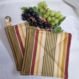 May include: Two green, red and yellow striped pot holders with a loop handle. The pot holders are folded in half and are sitting on a white surface. A bunch of green and black grapes are sitting on top of the pot holders.