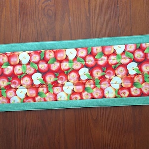 Peut inclure: Un chemin de table rectangulaire présentant un motif répétitif de pommes rouges, de feuilles vertes et de tranches de pommes blanches. Le tissu central à thème de pommes est bordé en haut et en bas par une bordure en tissu vert clair texturé.