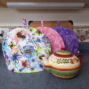 May include: A ceramic teapot with a green, red, and yellow striped design, next to three floral tea cosies. The tea cosies are white with colourful flowers, pink with a floral pattern, and purple with a floral pattern.