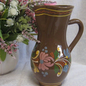 May include: A brown ceramic jug with a floral design in shades of pink, blue, green, and yellow. The jug has a wide mouth and a handle.