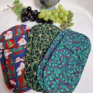 Peut inclure: Trois housses de bouteilles de vin sur le thème de Noël. L'une présente le Père Noël dans des carrés rouges et bleus, une autre a un motif floral vert et la troisième affiche un motif de houx et de baies bleu marine. Une tasse en céramique et des raisins sont en arrière-plan.