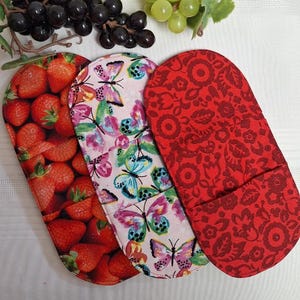 Peut inclure: Trois maniques de four avec différents motifs. L'une présente une impression de fraises, une autre des papillons colorés sur fond rose, et la troisième un motif floral rouge. Les maniques sont de forme ovale.