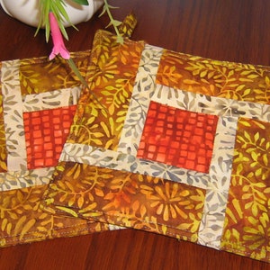 May include: Two brown and orange quilted pot holders with a white and grey border and a red square in the centre. The pot holders have a loop for hanging.