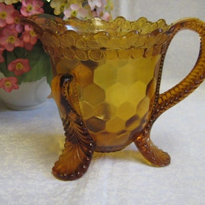 Peut inclure: Pichet en verre ambré avec un bord festonné et un motif floral. Le pichet a une poignée torsadée et trois pieds avec un motif floral.