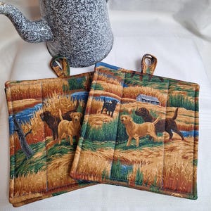 May include: Two brown and black quilted pot holders with a dog and field print. The print features brown and black dogs, tall grass, and a small wooden building.
