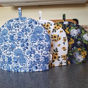 Peut inclure: Trois couvre-théières avec différents motifs floraux. Le premier est bleu et blanc avec un motif floral, le second est blanc avec des tournesols et le troisième est bleu marine avec des roses et des fleurs jaunes. Chaque couvre-théière a une poignée en boucle.