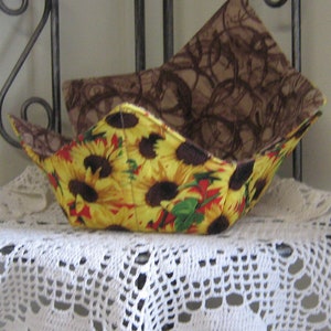 Peut inclure: Deux maniques à bols en tissu. La manique inférieure présente un motif de tournesols jaune vif avec des centres bruns et des feuilles vertes. La manique supérieure a un motif marron sur fond beige. Les maniques sont sur une étagère en dentelle blanche.