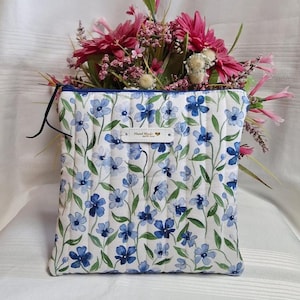 May include: A square quilted pouch featuring a white fabric base with a repeating pattern of blue flowers and green stems. The pouch has a dark blue zip closure and a small rectangular white label centred on the front with the text Hand Made.
