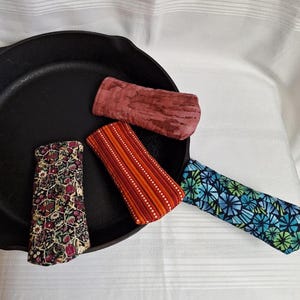 Peut inclure: Une poêle en fonte noire avec quatre manchons colorés. Les manchons présentent divers motifs, notamment floraux, géométriques et rayés. Les manchons sont dans les tons de rouge, orange, bleu et vert.