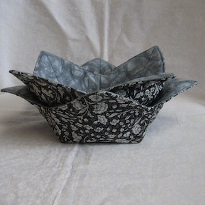 May include: Two fabric bowl cozies in a floral pattern. The outer fabric is black with white flowers and leaves. The inner fabric is a grey and white swirl pattern. The cozies are nested inside each other.