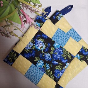May include: A yellow and blue quilted pot holder with a floral pattern. The pot holder has two blue fabric loops for hanging.