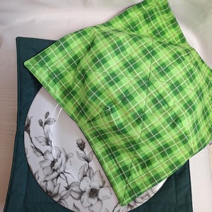 May include: A square fabric bowl cosy featuring a bright green and white plaid pattern on the top and a solid dark teal fabric on the bottom, with visible stitching lines forming a cross shape, holding a white plate with a black floral design.