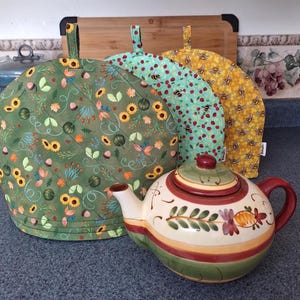 Peut inclure: Une théière en céramique avec un motif floral est devant trois couvre-thé matelassés. Les couvre-thé ont différents motifs : tournesols, coccinelles et abeilles. La théière est de couleur crème avec des accents rouges et verts.
