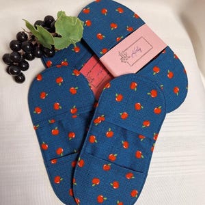 May include: Two blue fabric pot holders with a repeating red apple pattern and a small green leaf on each apple. A pink paper band with the text "Kathy" in cursive script wraps around the centre of the items. A cluster of dark purple grapes is visible.