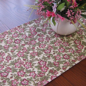 May include: A floral table runner with a cream background and a pink and green floral pattern. The runner is on a wooden table with a vase of pink flowers in the centre.