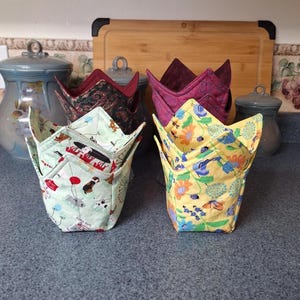 May include: A selection of handmade bowl cozies in different patterns. The cozies feature a light green fabric with dog and balloon prints, a yellow floral print, and burgundy and dark green patterned fabrics. These fabric bowl cozies are designed to keep food warm.