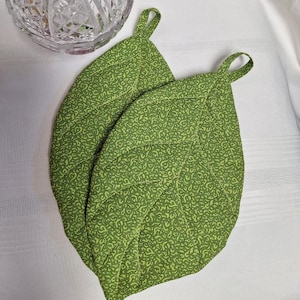 May include: Two leaf-shaped quilted pot holders in a vibrant green fabric featuring a small-scale, intricate tonal pattern. Each piece is constructed with visible stitching lines that mimic leaf veins and includes a small fabric loop for hanging.