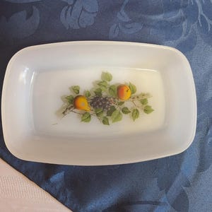 May include: A rectangular white milk glass dish featuring a central illustration of two yellow pears and a cluster of dark purple grapes surrounded by green leaves on a branch. The dish has rounded corners and a smooth, opaque finish.