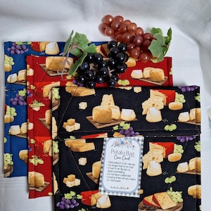 May include: Three fabric potato bags in blue, red, and black, each featuring a repeating print of cheese wedges and grapes. A white care card is attached to the black bag with text including "Potato Bag Care Card" and microwave instructions.