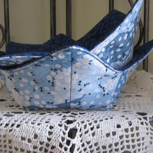 May include: Two fabric bowl cozies in a light blue floral pattern with white daisy-like flowers. The interior is a dark blue fabric. The cozies are stacked on a white lace doily, suggesting a kitchen or dining theme.
