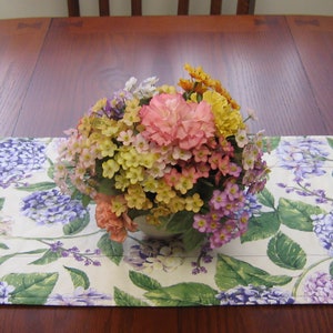 Peut inclure: Un arrangement floral de fleurs roses, jaunes et oranges est posé sur un chemin de table. Le chemin de table présente un motif d'hortensias violets et blancs avec des feuilles vertes sur fond crème. La table est en bois.